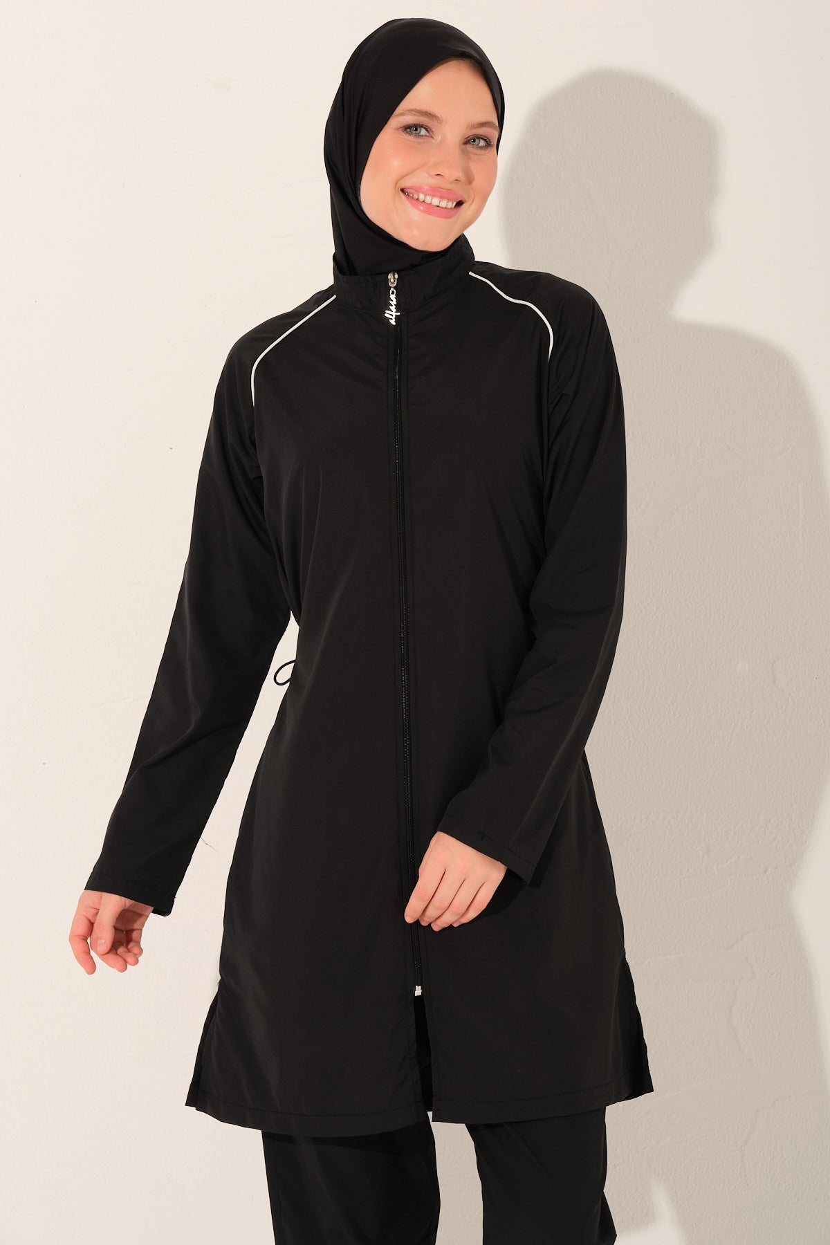Woman wearing a modest swimwear  black long-sleeve top with white zippers with white piping details on a plain background