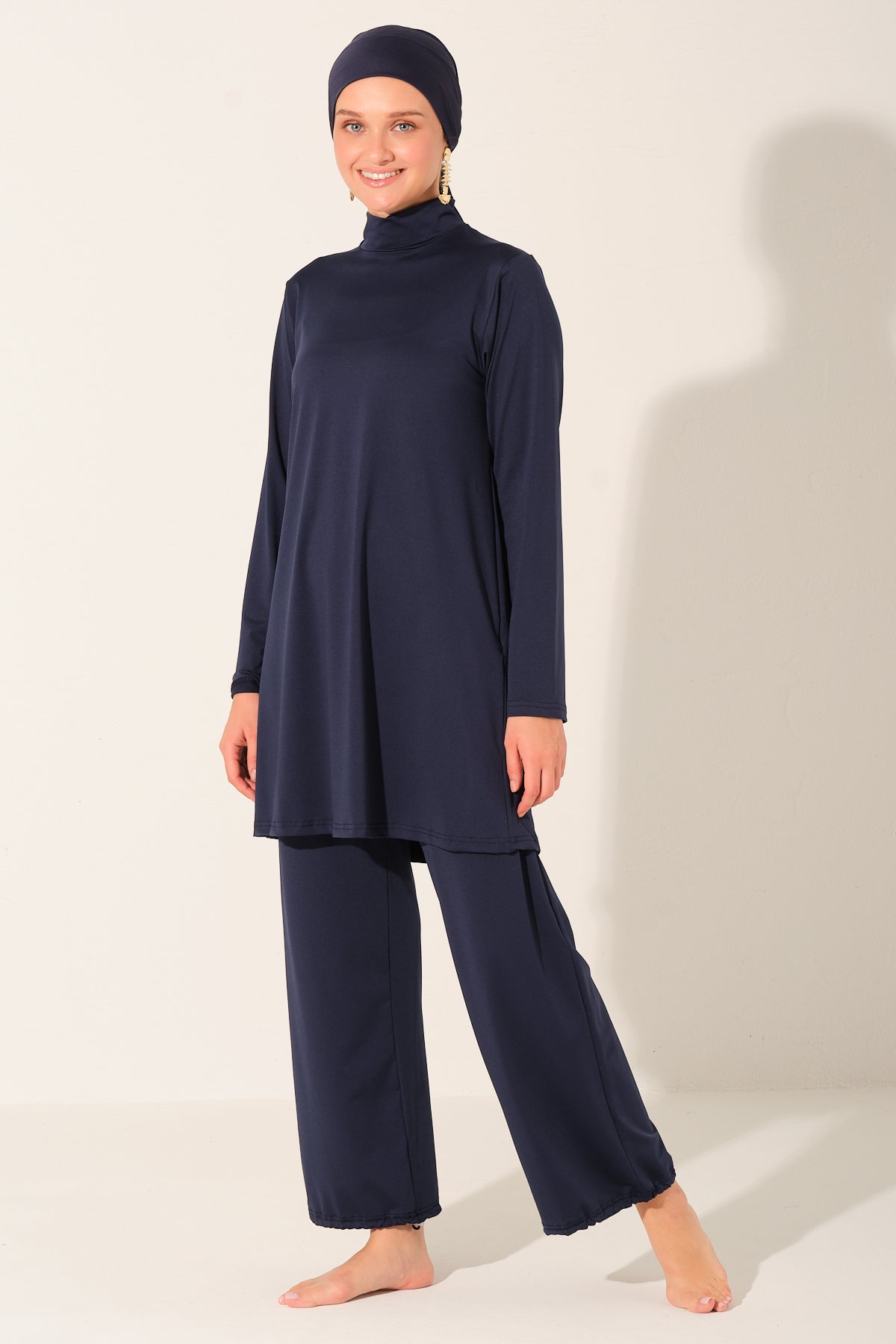 A woman modeling a navy blue two-piece modest outfit consisting of a long-sleeve mock-neck tunic and matching wide-leg pants, paired with a matching navy headscarf, against a plain off-white studio background.