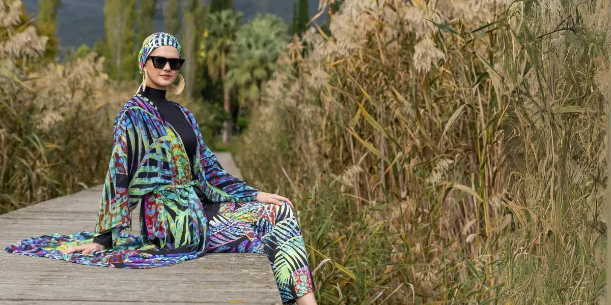 A woman wearing a vibrant, multicolored tropical print kimono and matching headwrap with cat-eye sunglasses, posing on a wooden boardwalk surrounded by tall, golden reeds.