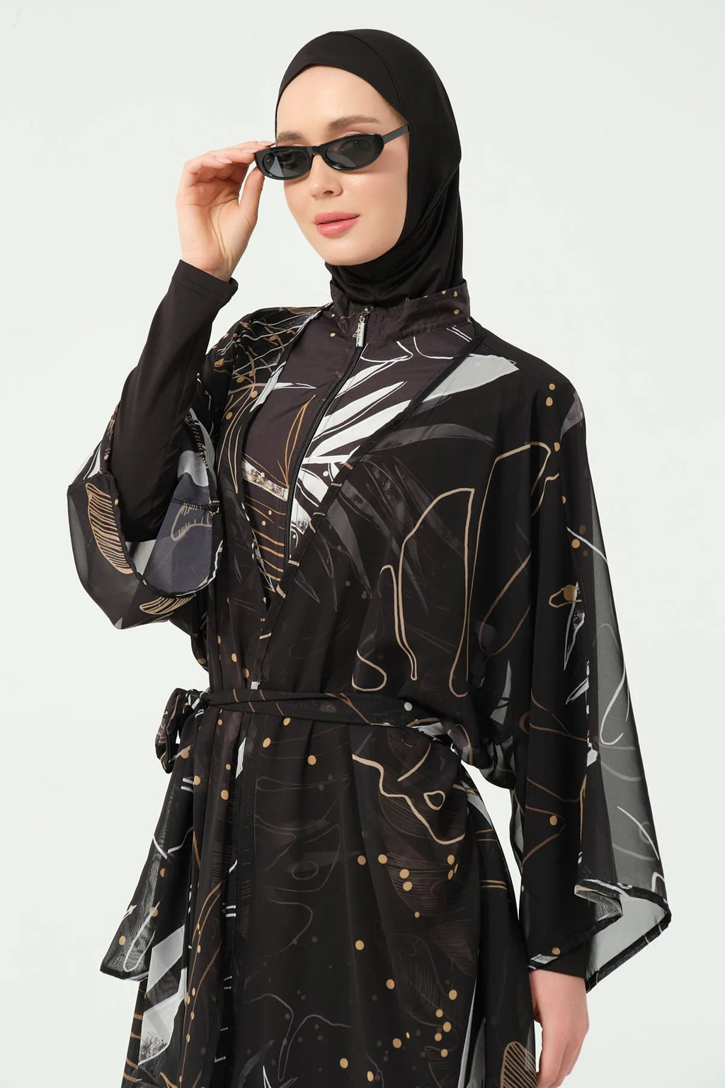 Woman wearing a black and gold patterned outfit with a hijab and sunglasses on a white background