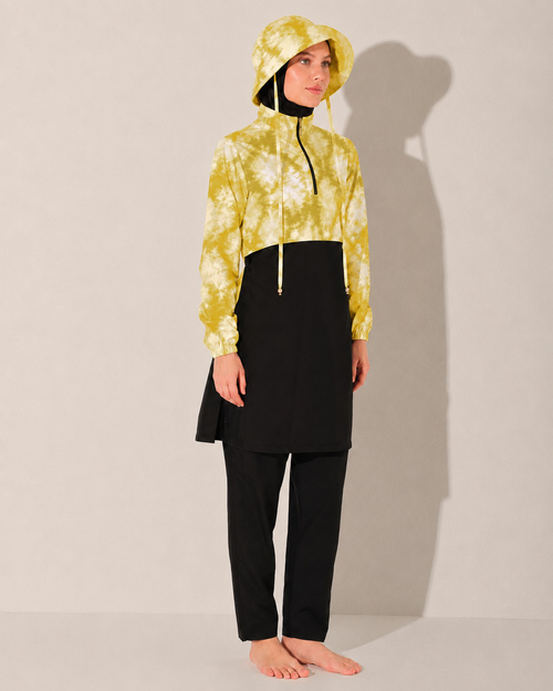 A model stands barefoot against a light beige studio wall, wearing a hooded long-sleeve top with a yellow and white tie-dye pattern on the upper half and black on the lower half modest swimwear, paired with black pants.