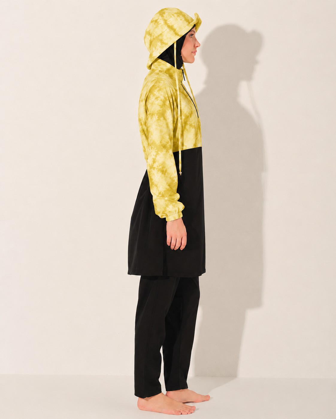 A model stands in profile wearing a burkini long-sleeve dress or tunic with a yellow tie-dye upper section and hood, and a solid black lower section and black pants, against a plain off-white studio background with a strong shadow.