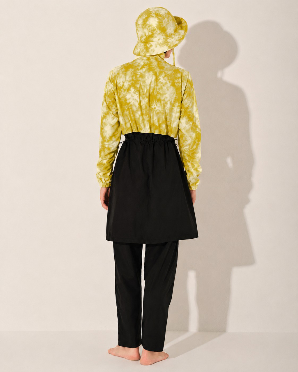 A model is shown from behind wearing a yellow and white tie-dye long-sleeve shirt burkini , a black high-waisted skirt, and black pants, standing against a plain off-white wall with a strong shadow.
