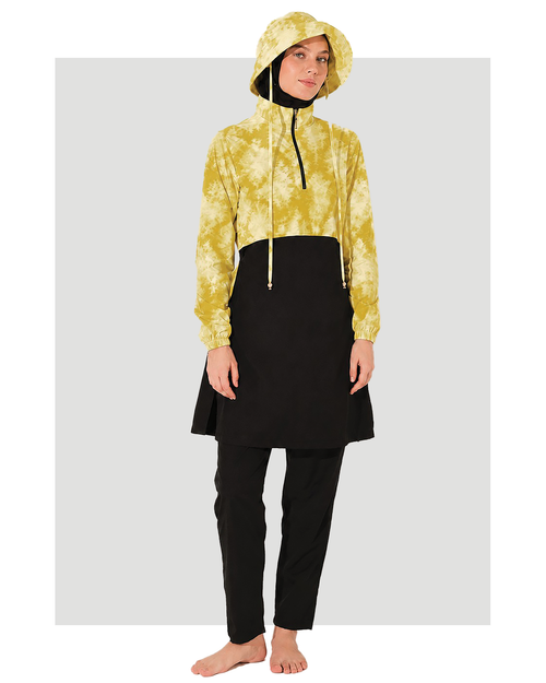A model wearing a yellow and white tie-dye cropped hooded sweatshirt with black drawstrings, layered over a black tunic, black pants, and a black head covering burkini, standing barefoot against a light gray background.