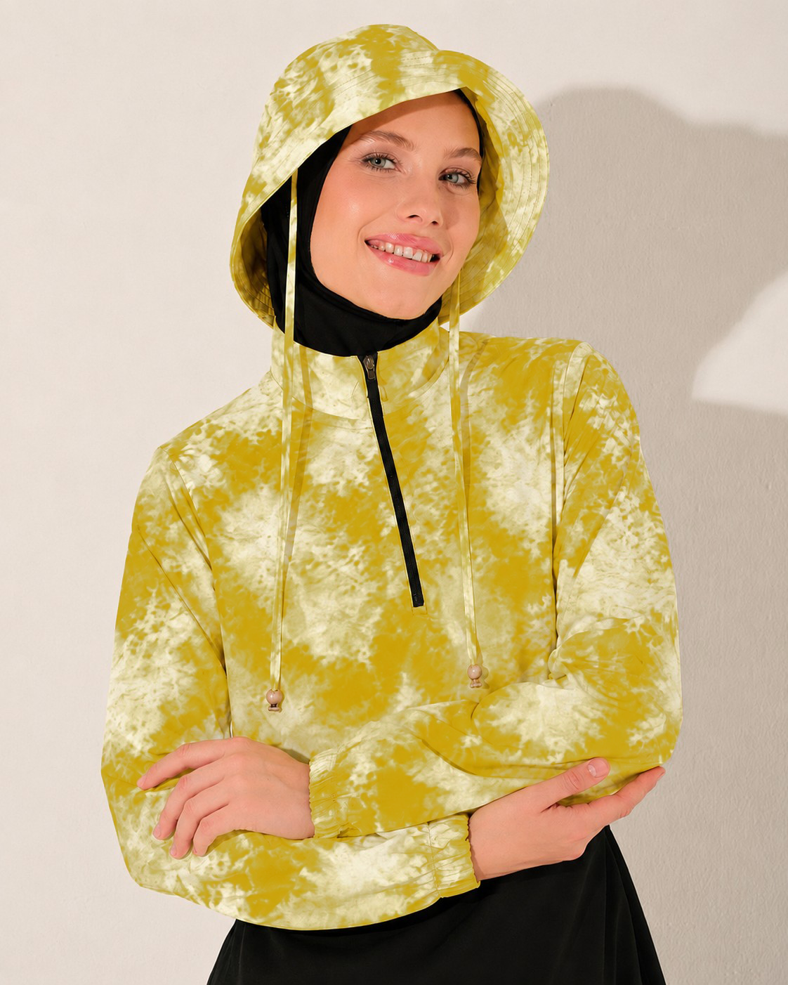 A woman wearing a yellow and white tie-dye hooded sweatshirt with a black inner neck gaiter and black bottoms burkini, posing against a neutral light gray wall.
