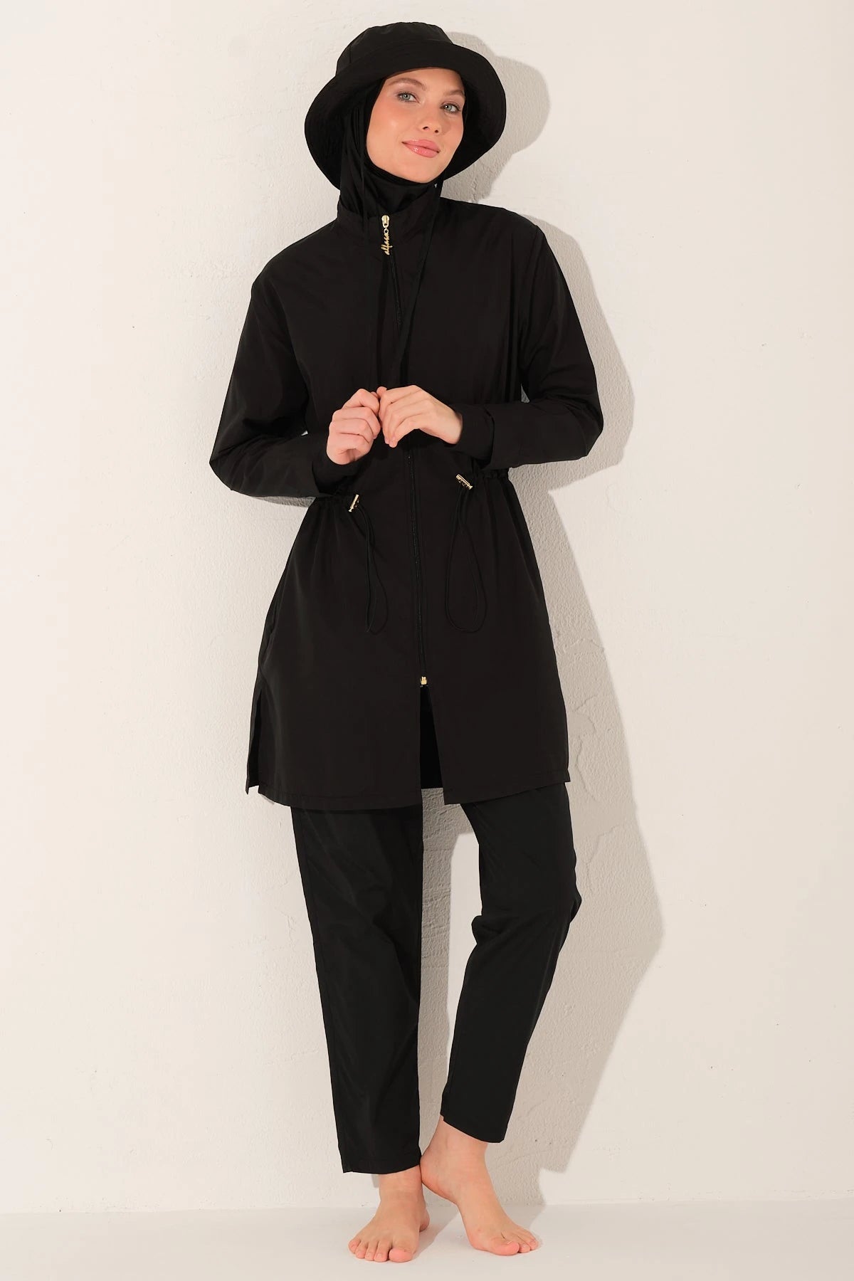 Woman in black burkini set with bucket hat, hands clasped in front.