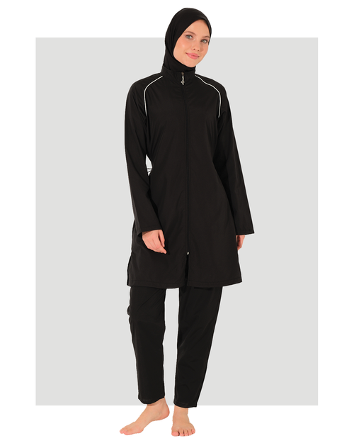 A woman modeling a black modest swimwear set consisting of a long-sleeve high-neck swimsuit with white piping details, matching black swim pants, and a black hijab, set against a light gray background.