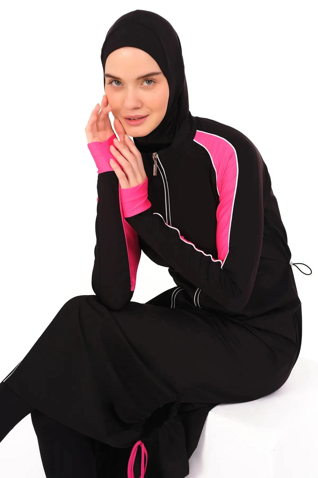Model seated in black burkini with pink sleeves, hands near face.