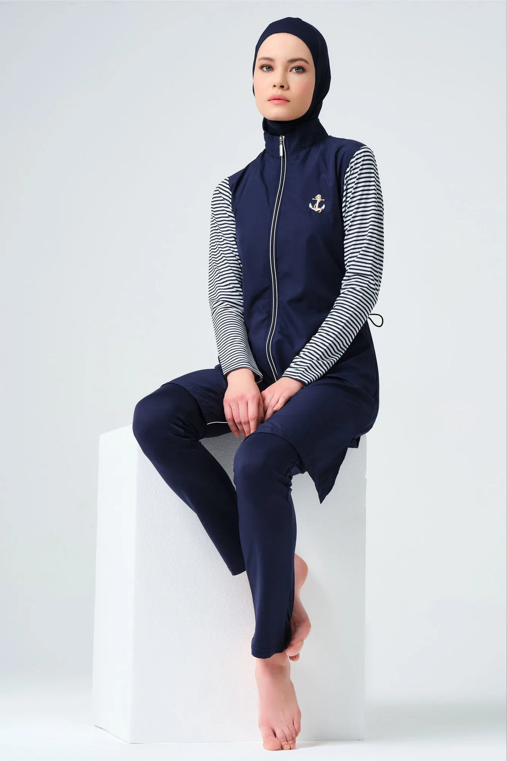 Model sitting on a white block in navy burkini with striped sleeves.
