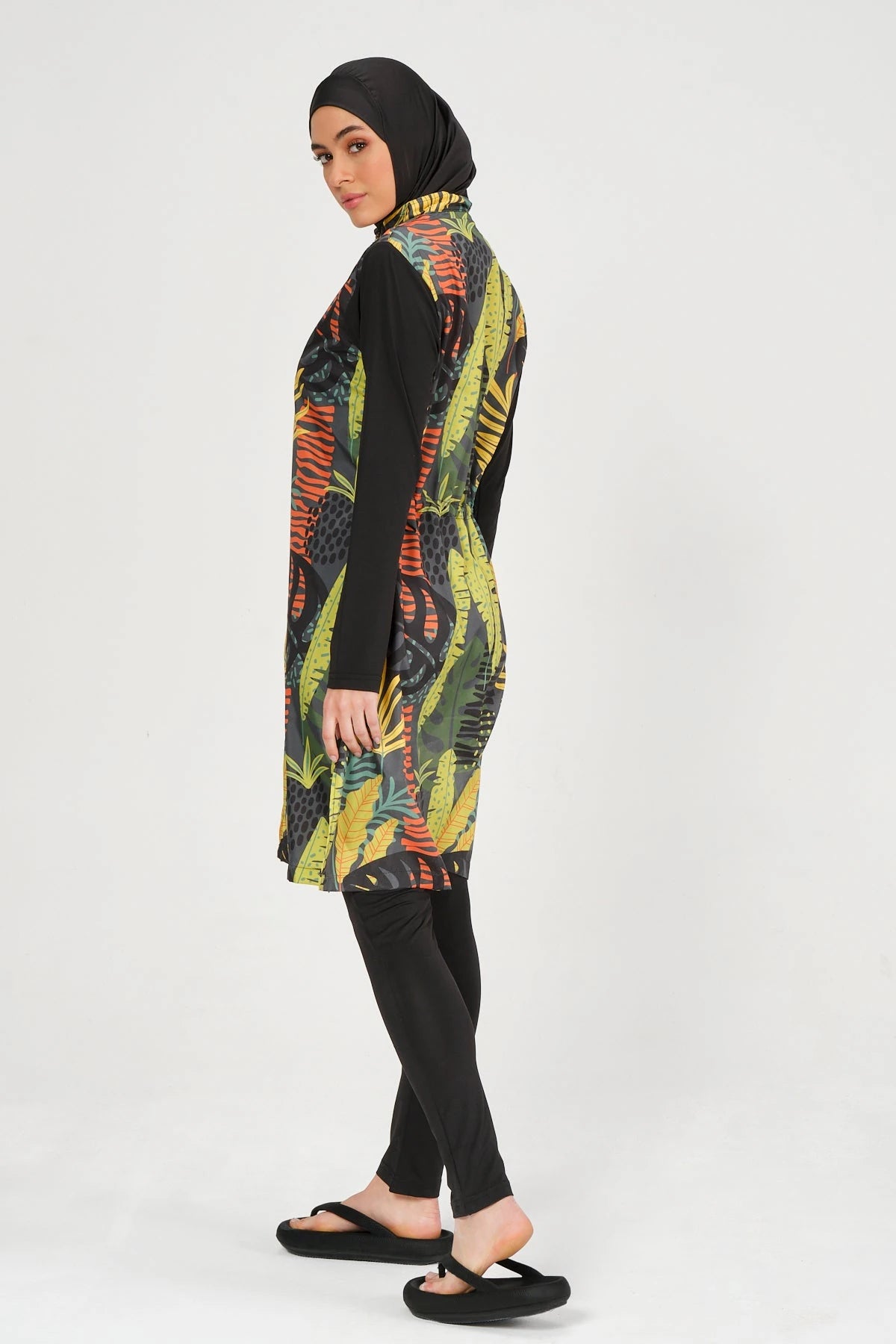 Woman in green tropical print burkini with black leggings and hijab, side view.