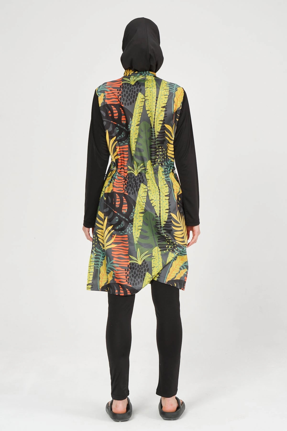 Woman in green tropical print burkini with black leggings and hijab, back view.
