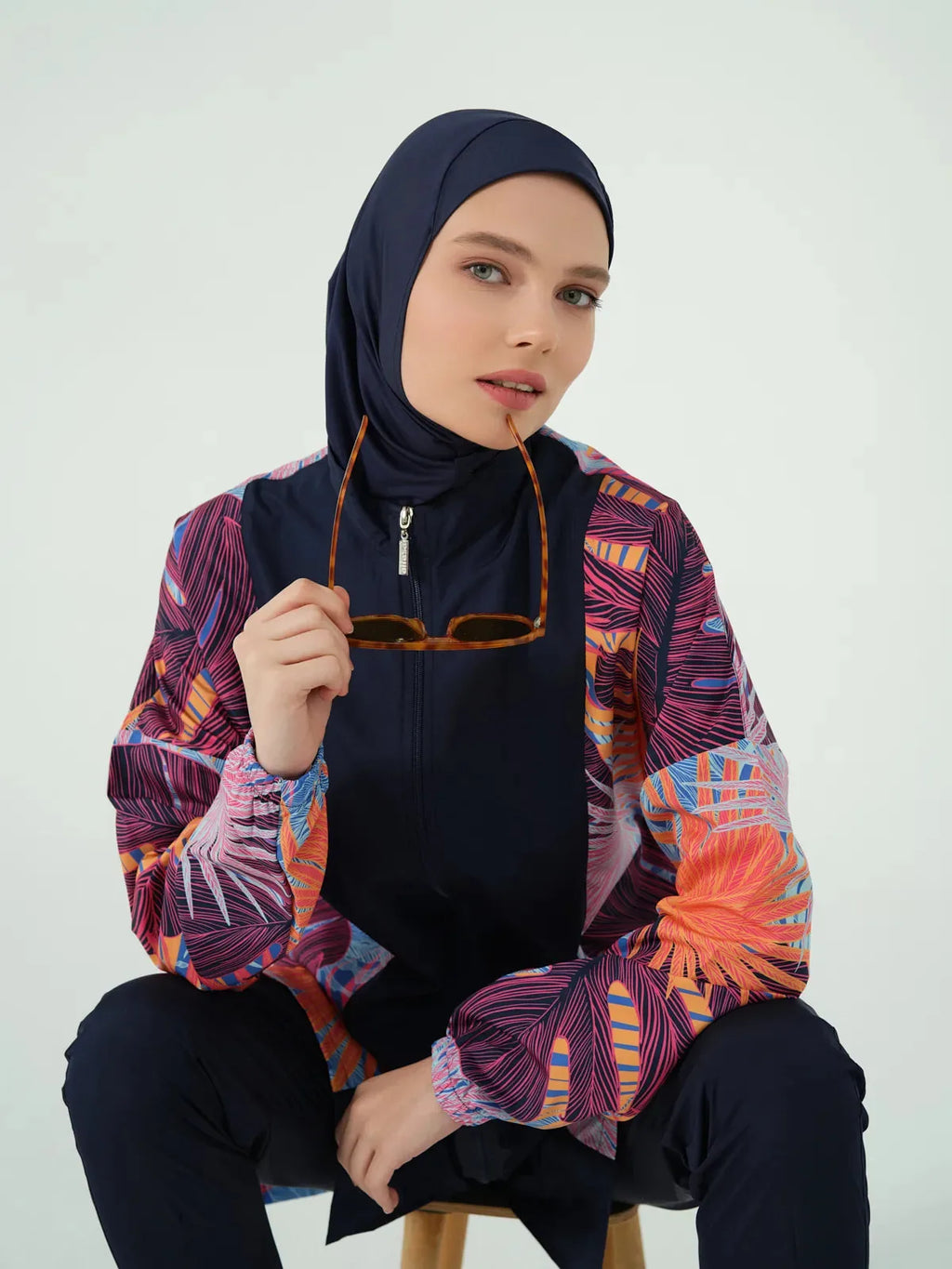 Woman sitting in modest swimwear, navy hijab, holding sunglasses.