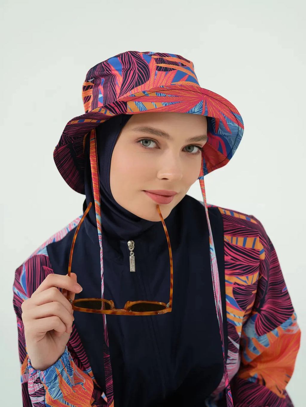 Close-up of woman in navy hijab, tropical print hat, holding sunglasses.