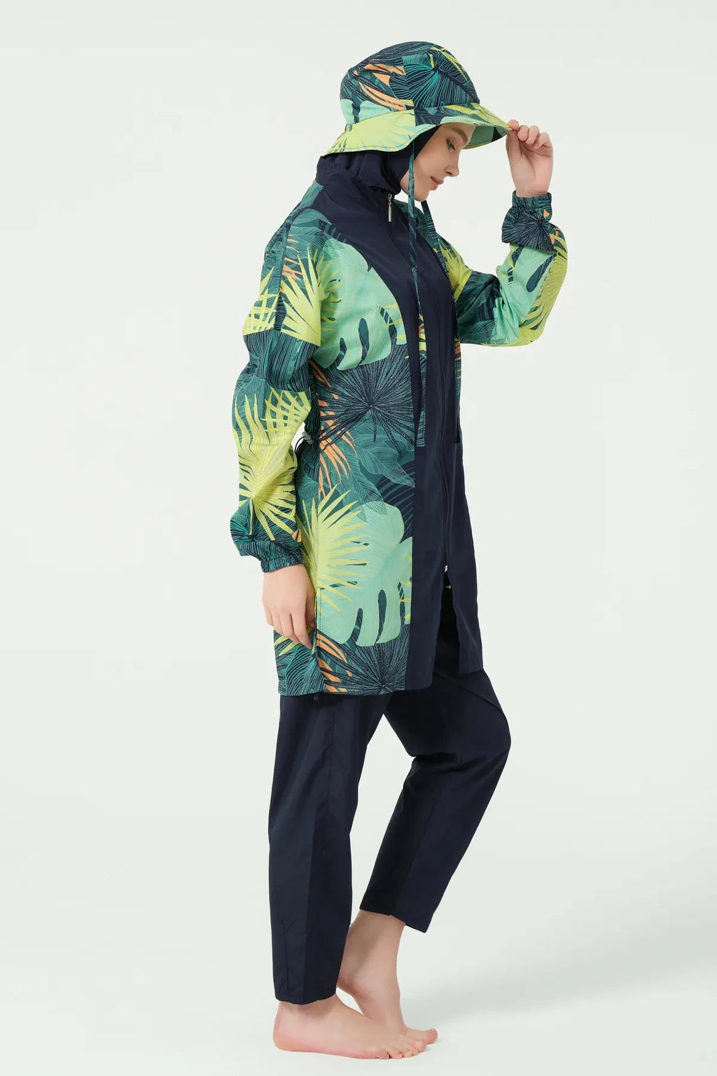Model in tropical green burkini set adjusting matching bucket hat, side view.