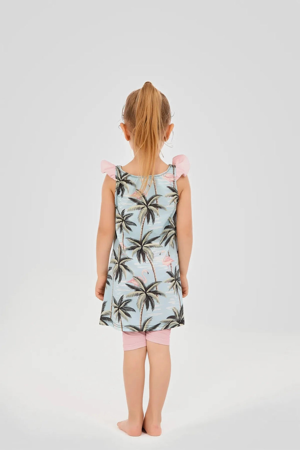 Rear view of a young girl in a light blue flamingo and palm tree print modest swim dress.