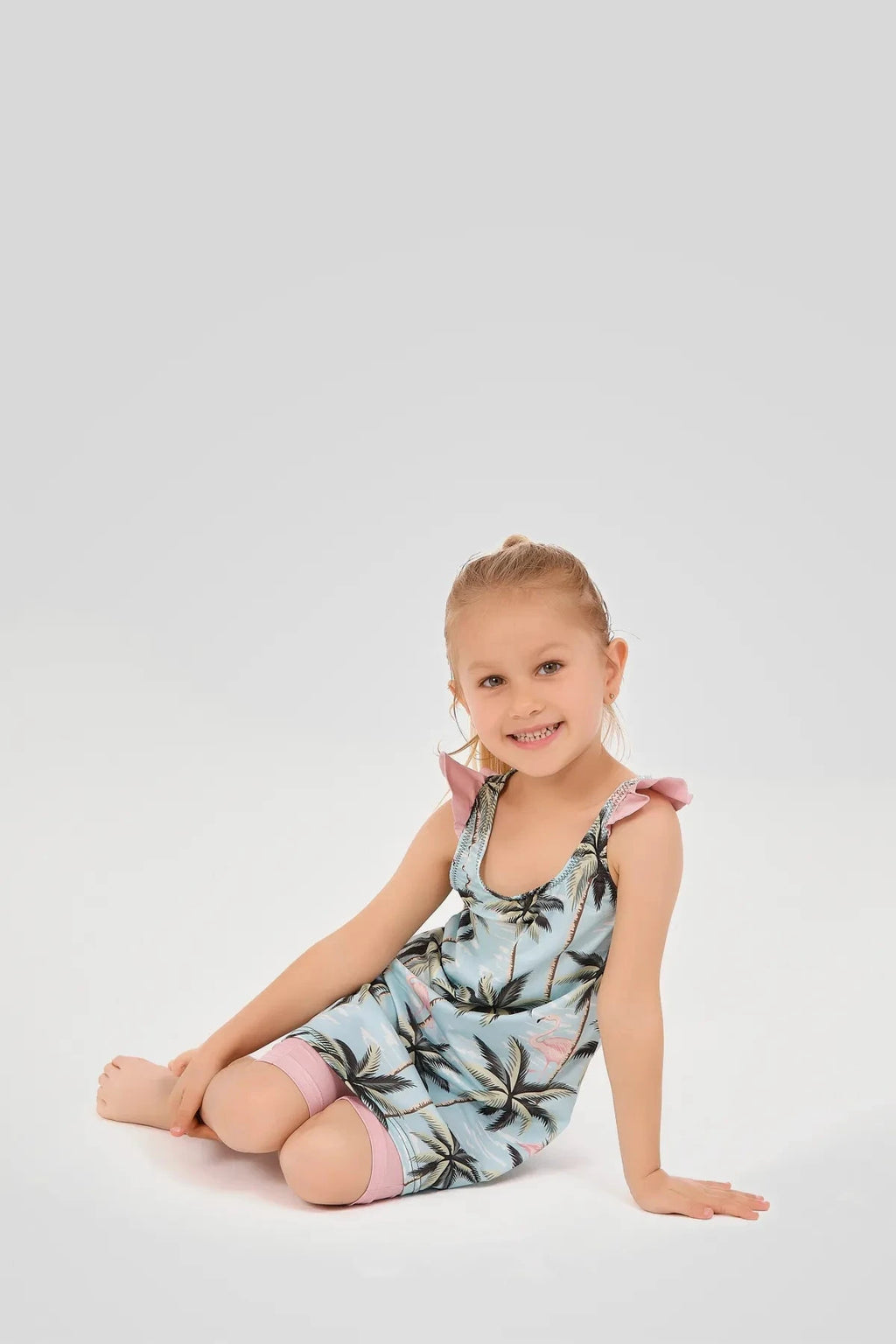 Young girl sitting down in a light blue flamingo and palm tree print modest swim dress.