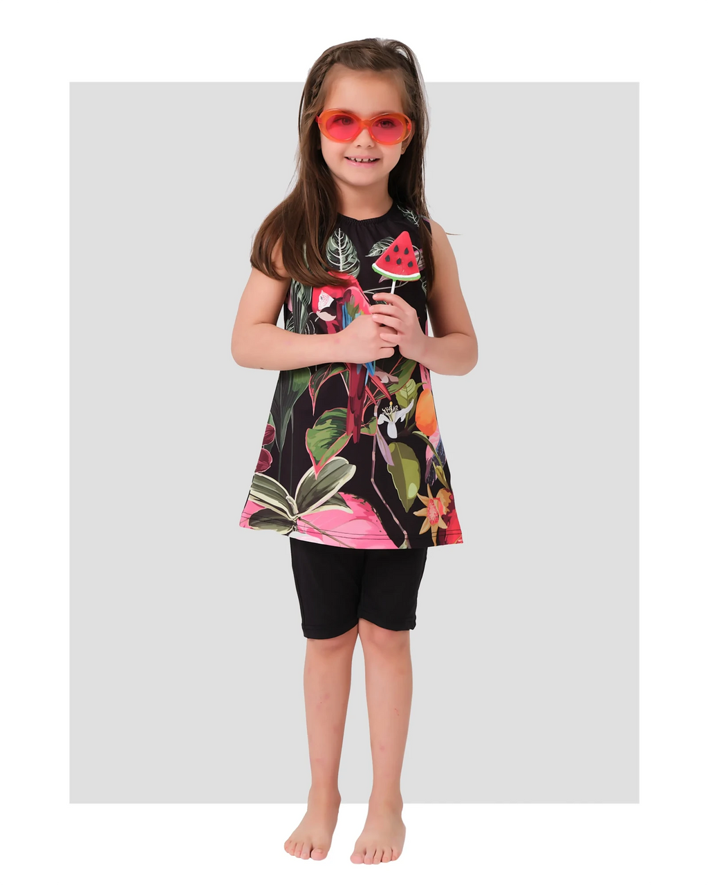 Young girl in floral print swim dress and black shorts, holding a watermelon lollipop.