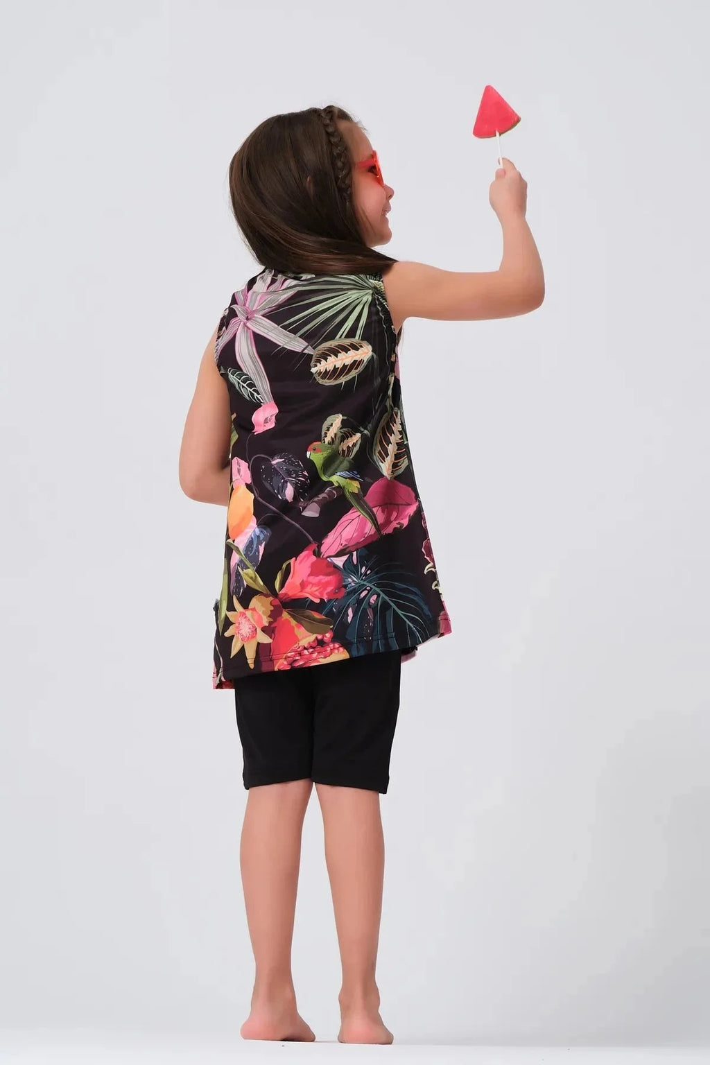 Back view of a girl in a floral print swim dress and black shorts, holding a watermelon lollipop.