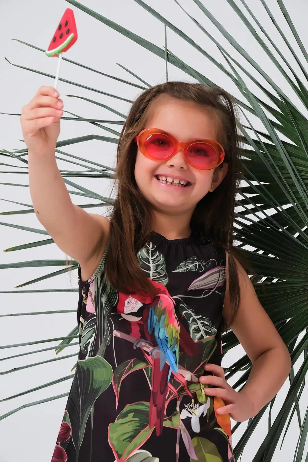 Close-up of a girl in a tropical print swim dress, wearing sunglasses and holding a lollipop.