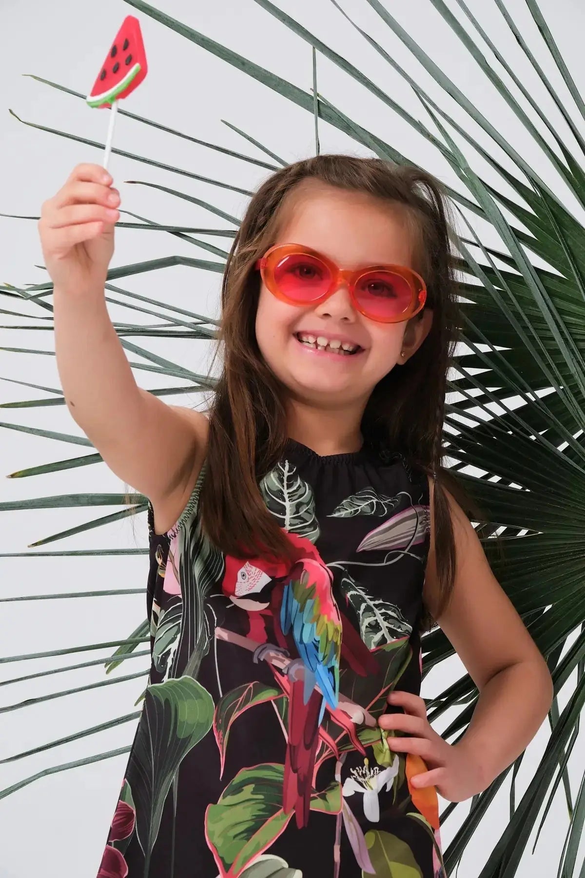 Close-up of a girl in a tropical print swim dress, wearing sunglasses and holding a lollipop.