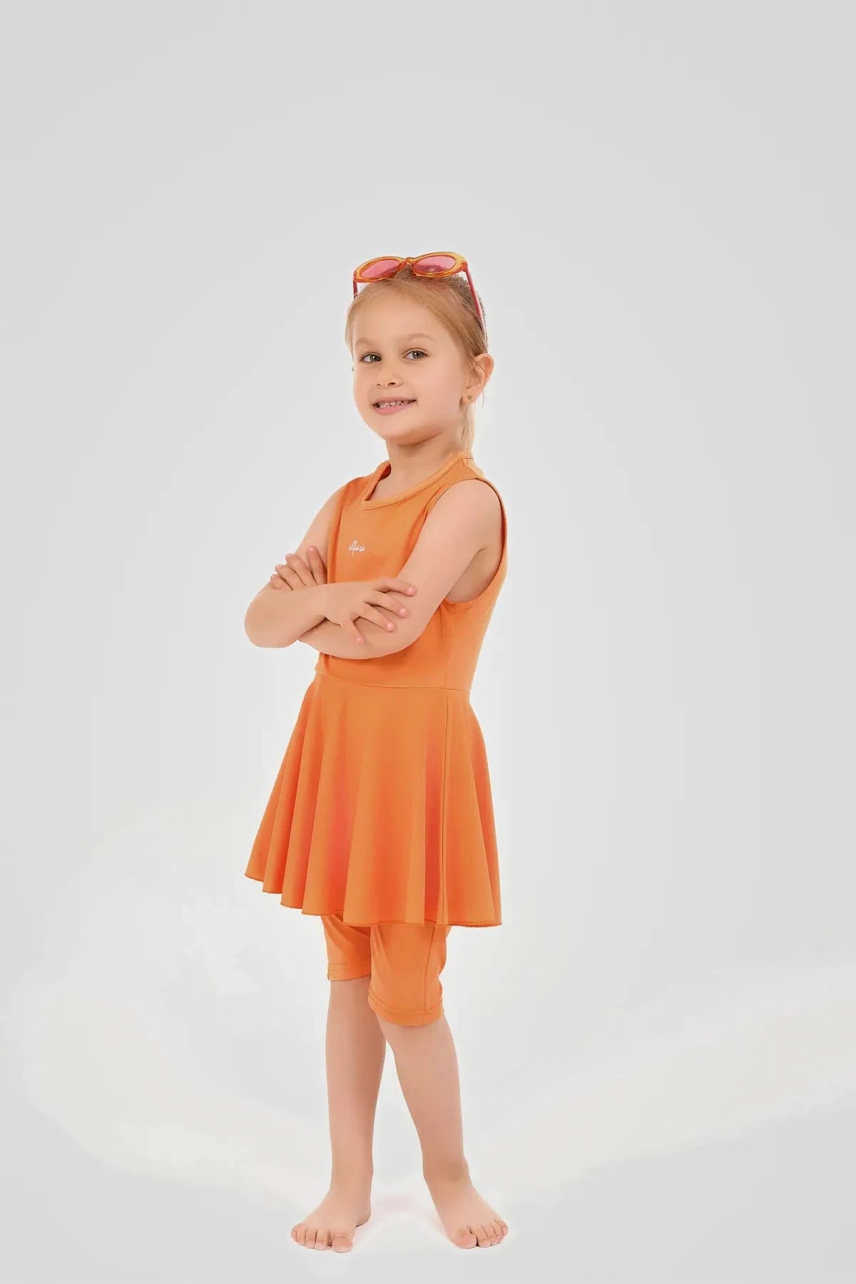 Young girl in orange modest swim dress and leggings, arms crossed.