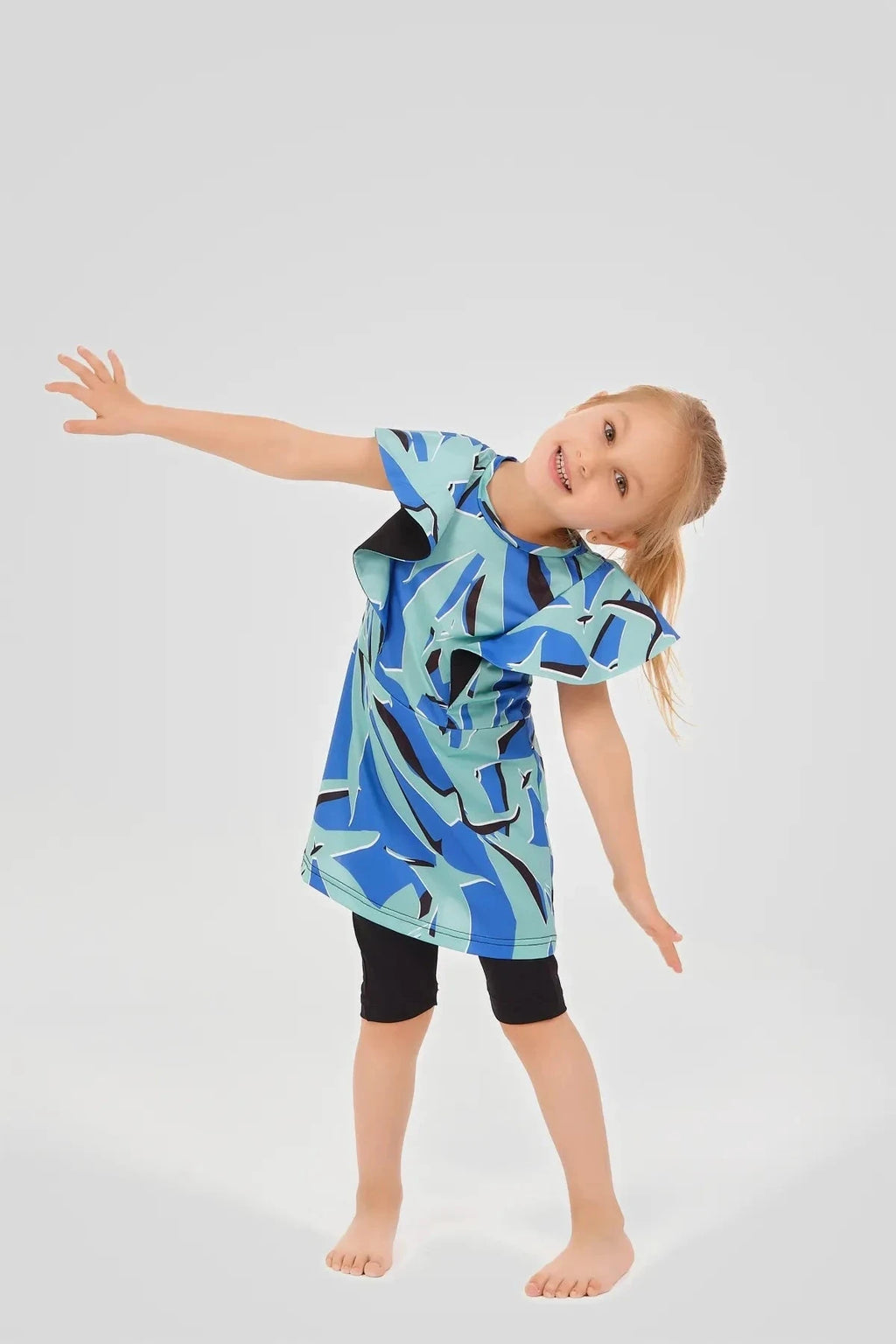 Child wearing blue abstract print burkini dress with black shorts, arms outstretched.