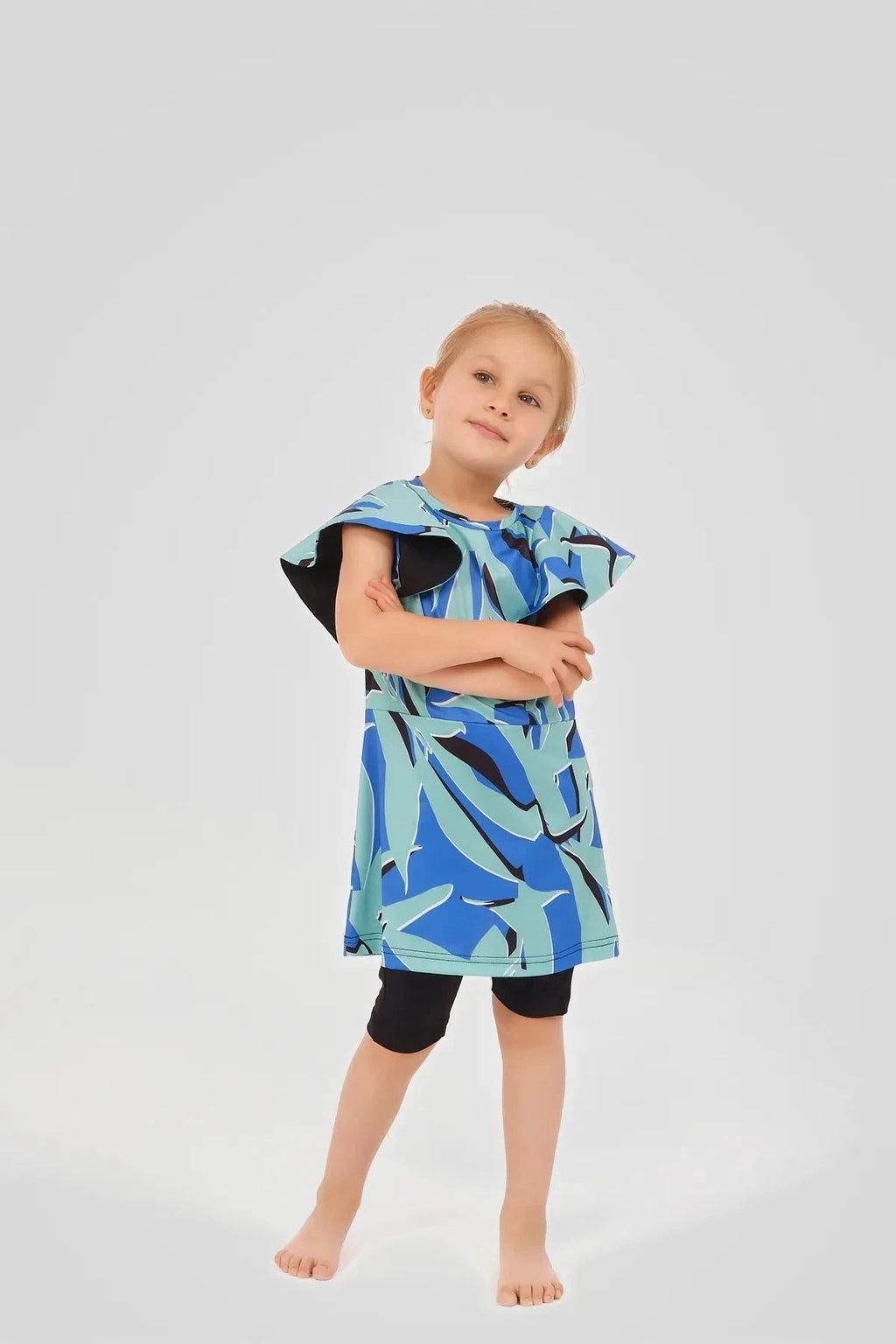 Child in blue abstract print burkini dress with black shorts, arms crossed.