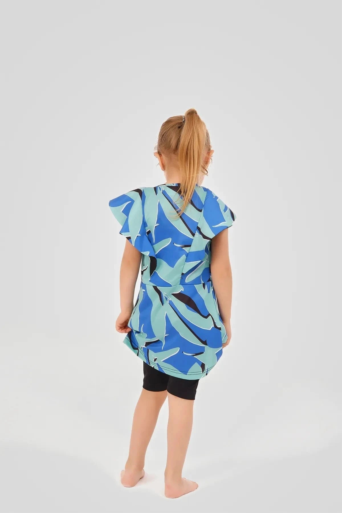 Child wearing blue abstract print burkini dress with black shorts, back view.
