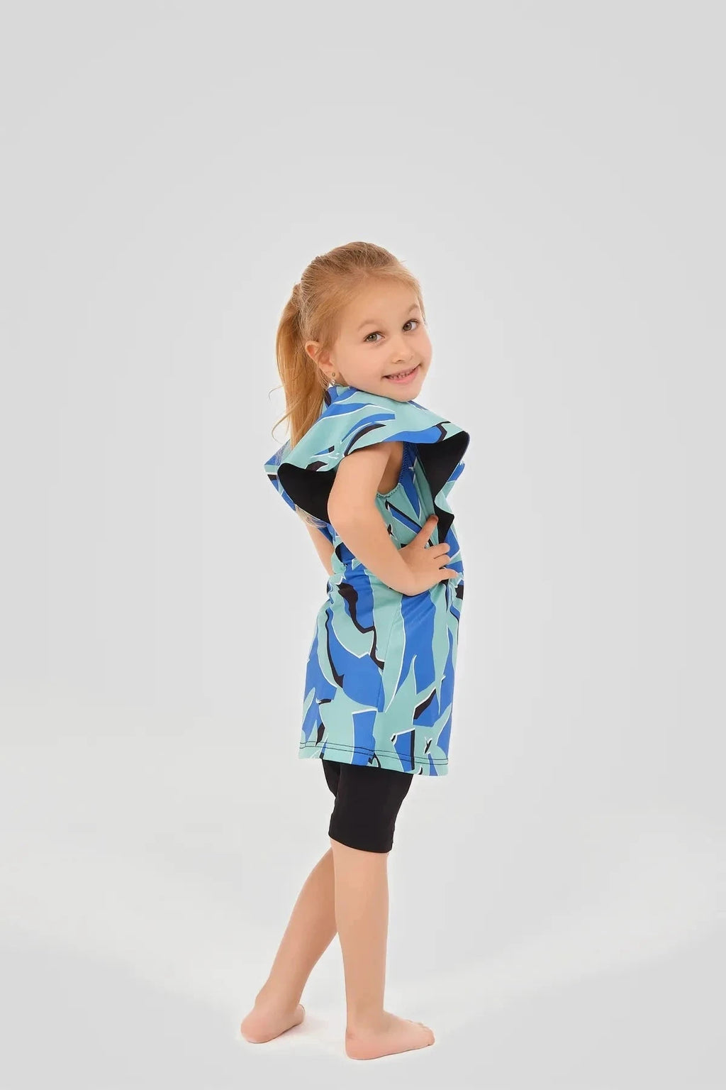 Child wearing blue abstract print burkini dress with black shorts, hands on hips.