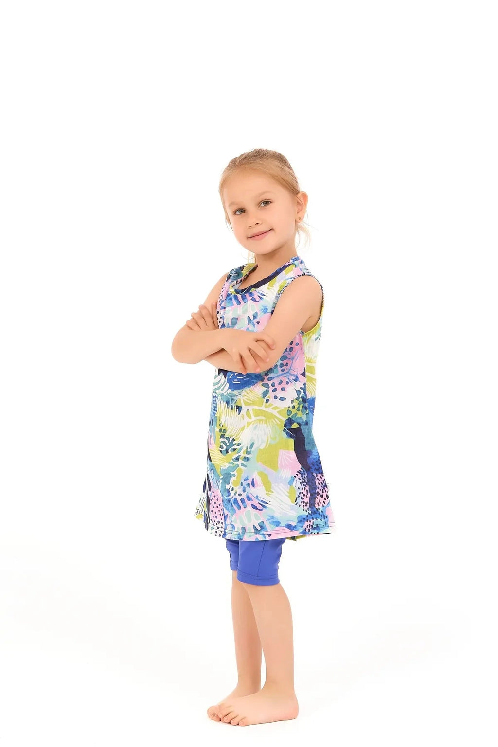Young girl in a floral print burkini dress and blue leggings, arms crossed.