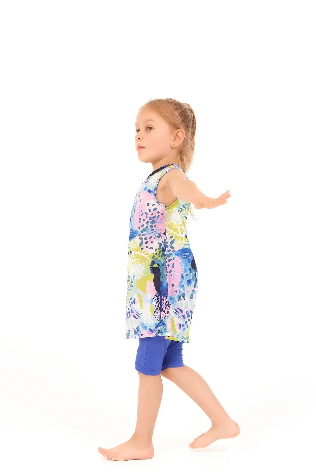 Young girl in a floral print burkini dress and blue leggings, side view.