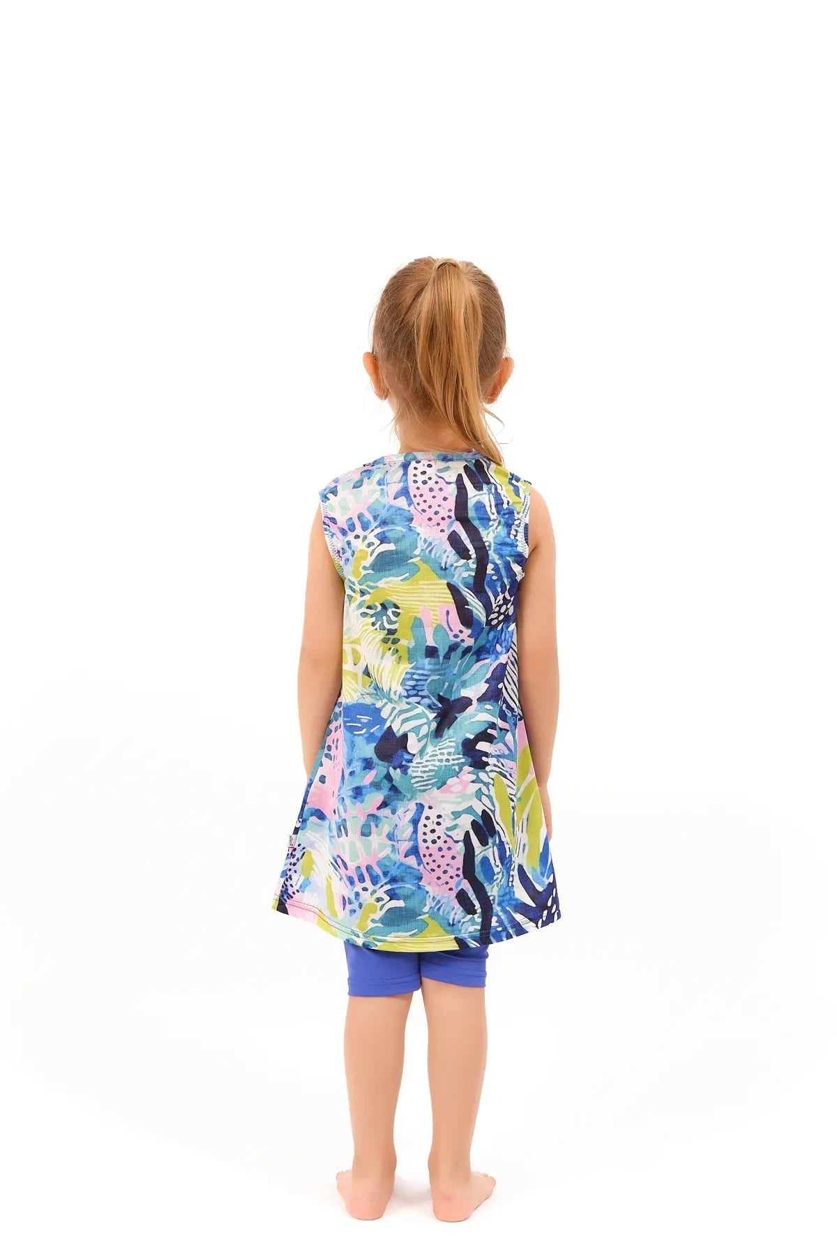 Young girl in a floral print burkini dress and blue leggings, back view.